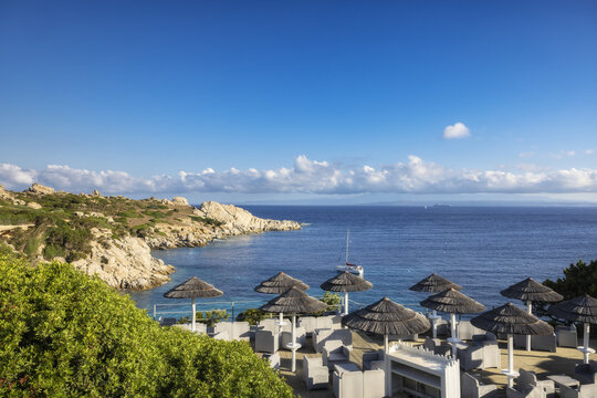 View of sun-kissed straw umbrellas dotting the serene coast, where turquoise waters meet rugged, rocky cliffs under a vast, azure sky, Capo Testa, Sardegna, Italy.