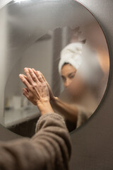 Naklejka premium Woman wiping foggy bathroom mirror with hands after shower, soft reflection through steamed glass, towel on head, morning hygiene ritual, self care wellness concept at home