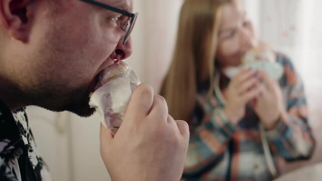 Closeup and side view of caucasian bearded man with glasses that bites and eats easter cake. Young blurred woman in plaid shirt sits and savors. Eating homemade baked goods on a holiday weekend