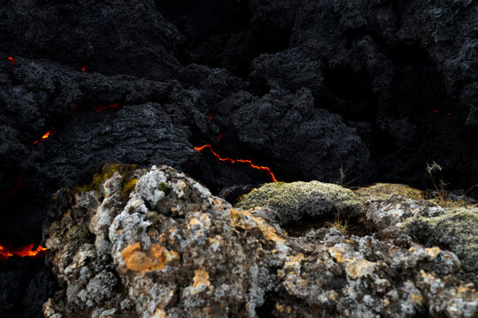View of molten lava snaking through jagged, black volcanic rock, juxtaposed with mossy, weathered stones, a stark reminder of nature's power, Grindavik, Iceland.