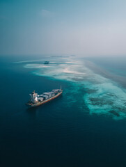 aerial view of a lone cargo ship in the center of a vast, calm turquoise ocean. The water is crystal clear, showing subtle variations in depth and light. The ship is loaded with grey and white 