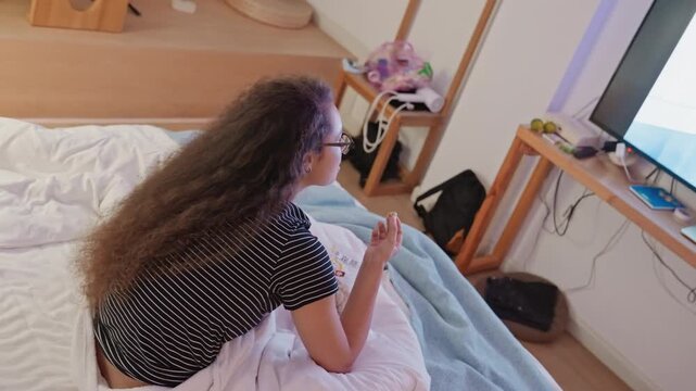 Woman using remote in hotel room, reaching toward TV while sitting on bed with blanket pulled up, bedside table and soft lamp frame focused streaming session and quiet evening interaction
