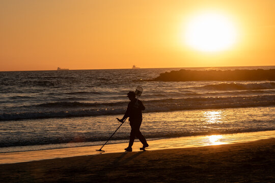 View of a lone figure silhouetted against the vibrant orange glow of the setting sun while scanning the sandy shore with a metal detector, Newport Beach, California, United States.