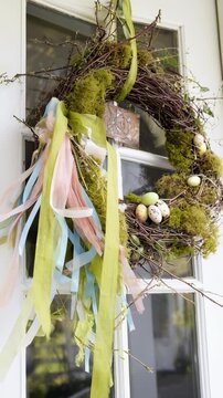 An Easter wreath made of twigs, moss, and small quail eggs hanging on a door, ribbons fluttering in a breeze, welcoming vibe