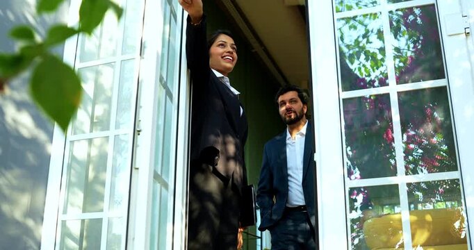 Indian woman real estate agent shows view from window to a prospective young married couple buyer during a home walkthrough while they admire the outside greenery and natural sunlight.
