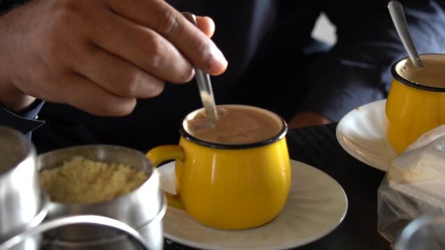 Person adding raw sugar and stirring hot tea in a yellow mug
