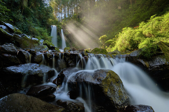 View of sunlight piercing through the lush canopy onto the cascading waters of a double waterfall flowing over moss-covered rocks, Magelang Regency, Central Java, Indonesia.
