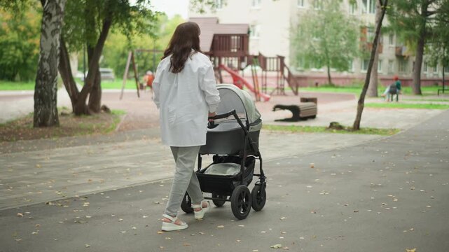Woman pushing stroller through park, quiet playground in background, soft autumn light, leafy trees, paved path, relaxed pace, casual sneakers, modern stroller, peaceful parenting moment, emphasis