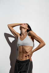 Athletic adult woman with defined abdominal muscles posing in sunlight against white wall,...