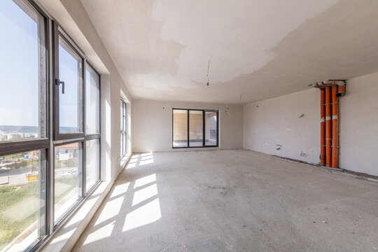 Interior of an empty room under construction in a new apartment. Unfinished space with raw concrete walls, floor, and a large window. Renovation and real estate concept.