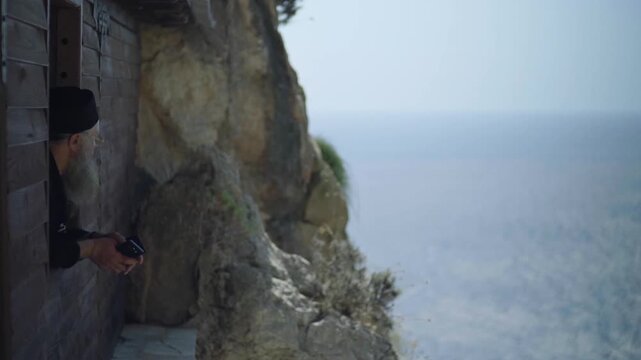 Elderly bearded monk in black cassock taking pictures of the sea from a monastery window