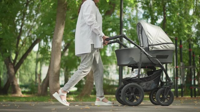 Fullframe woman pushing stroller through park caucasian mother in relaxed blazer and trousers walking past bench and birch trees soft daylight composed pace reflective mood urban park lifestyle decree
