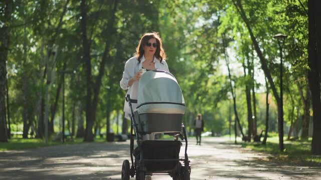 White mother pausing with stroller on treelined path, thoughtful gaze into distance, soft sunlight and shade create reflective calm, small moment of parenting lifestyle decree and solitude.