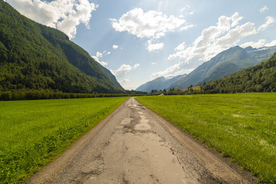 View of a long, cracked road stretches between vibrant green fields towards towering mountains under a sky dappled with fluffy white clouds, Supphellebreen, Norway.