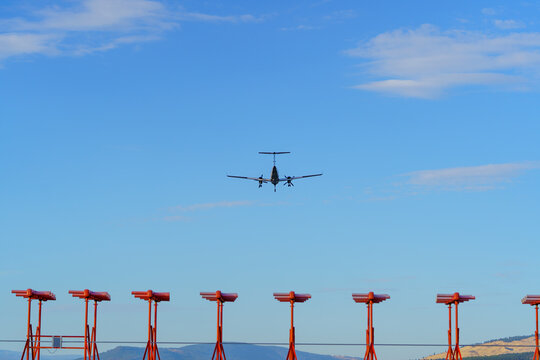 A twin-engine aircraft approaches for landing above a line of red airport approach lighting structures against a clear blue sky. Final approach phase of flight with infrastructure guiding the landing.