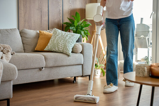 Woman in jeans vacuuming floor with cordless vacuum cleaner in living room beside sofa