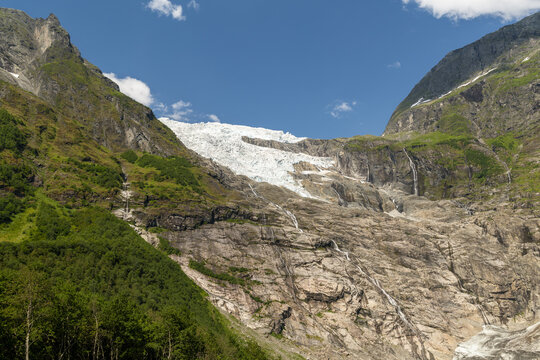 View of boyabreen glacier's icy tongue clinging to the rugged mountain face, contrasting with the vibrant green forest below, Fjaerland, Norway.