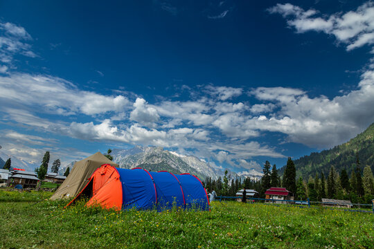 View of vivid tents nestled in a lush green meadow beneath a dramatic sky of fluffy clouds, with snow-capped mountains in the distance, Arang Kel, Pakistan.