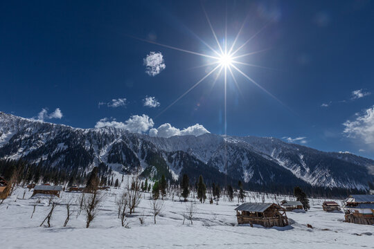 View of rustic wooden huts nestled in the snowy expanse beneath towering mountains touched by the sun's radiant glow, Arang Kel, Azad Kashmir, Pakistan.