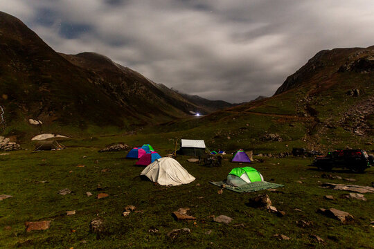 View of colorful tents dotting the lush green field against the backdrop of imposing mountains under a dramatic, cloudy sky, Lawat Bala, Pakistan.