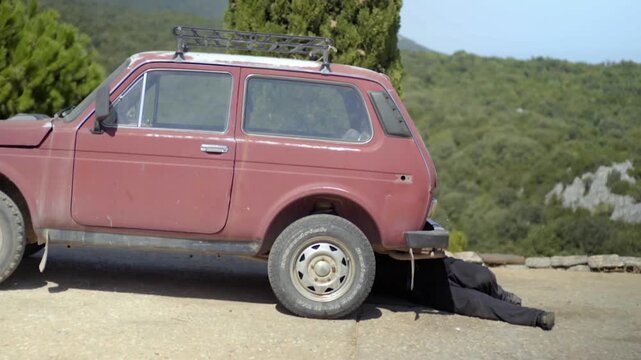 Man in black cassock lying under a broken down vehicle, trying to repair it on a rural road