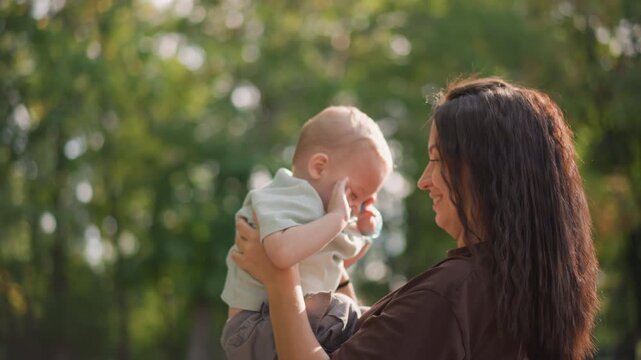 Mother lifts baby in sunshine, playful interaction in leafy park, pacifier visible, laughter and rhythmic motion, candid lifestyle moment, family decree of active caregiving and joy