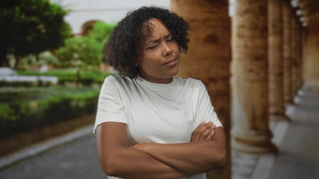 Young woman crosses arms over chest and tilts head while standing by building columns overlooking park greenery; rejection.
