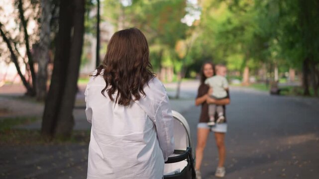 Park stroller meeting between two women greeting baby warm afternoon exchange on leafy path, woman approaches carrying infant while other returns to stroller, candid neighborly moment, genuine