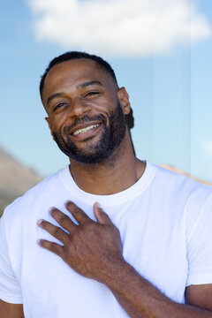 African American man posing outside under blue sky, wearing white tee, right hand on chest, smiling