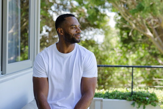 Mature African American man sitting on balcony ledge with railing, wearing white tee, gazing out