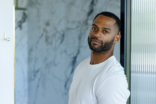 Mature adult African man leaning on ribbed glass in marble hallway wearing white tee, copy space