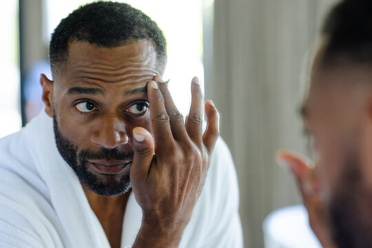 Middle-aged African American man touching under eye with fingers in white robe at dressing mirror