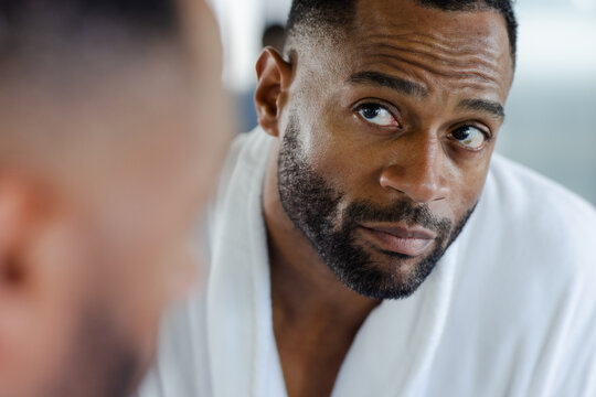 Mature man wearing white bathrobe gazing at reflection in large bathroom mirror closely