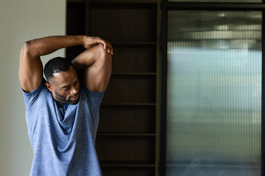 Mature man stretching triceps in room wearing heather tee by shelf and glass panel, copy space
