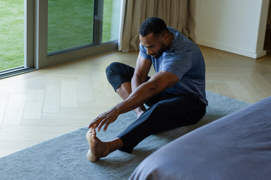Mature African American male stretching on gray rug in bedroom near sliding door wearing blue shirt