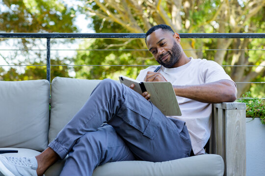 African American man sitting on wooden terrace sofa writing in notebook with pen phone nearby