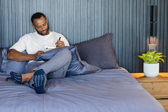 Mature man reclining against stacked pillows on bed, using tablet with stylus, copy space