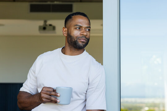Mature African American man standing at metal frame glass door holding ribbed mug gazing right