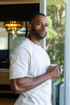Mature adult African male standing near sliding glass door holding mug by chandelier and gazing out