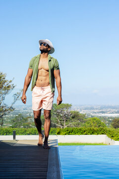 Man is walking barefoot along wooden pool deck wearing white wide brim hat and sunglasses