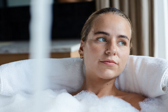 Female reclining in bathtub soaking in foam, resting head on rolled towel by bathroom faucet