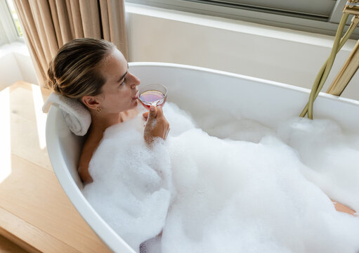 Adult female reclining in standalone tub with bubbles, holding stemmed glass, resting head on towel