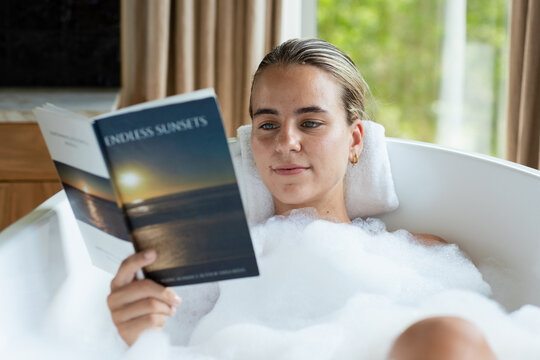 Woman reclining in bathtub reading ENDLESS SUNSETS amid thick bubbles, rolled towel, window light