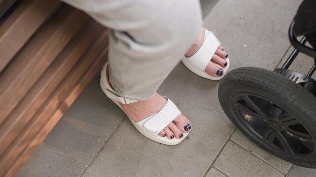 Bench scene sandals stroller closeup showing white braided sandals and stroller wheel on city pavement during quiet parenting lifestyle decree moment, casual beige trousers, black toenail polish,