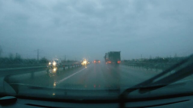 POV shot from inside a car driving on a wet highway in bad weather. A large truck is visible. Gloomy, overcast sky and red taillights create a somber mood. Focus on travel and poor visibility