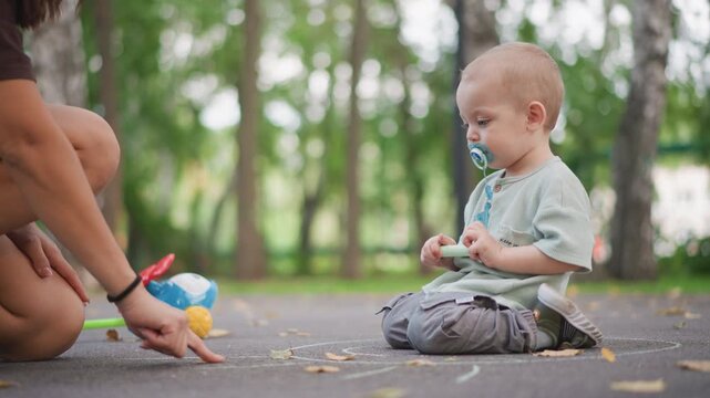 White toddler in park with pacifier, caregiver pointing to ground while child sits by toy and scattered leaves, gentle guidance and early routine capturing family decree and lifestyle moment