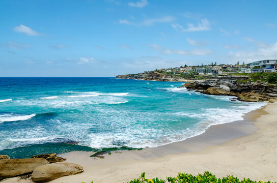 Tamarama Beach in Bondi, Sydney, Australia, featuring sandy shores and rolling waves. The ocean is a vibrant turquoise color under a clear blue sky dotted with clouds.