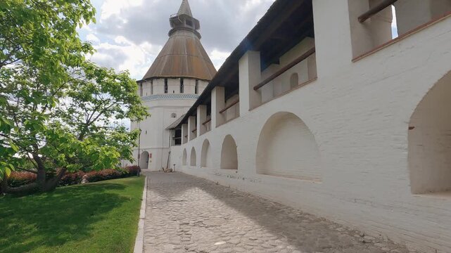 A timelapse of an ancient fortress from a low angle. A paved path leads along a whitewashed brick wall with arches to a large wooden tower. On a quiet day, landscape is framed by lush green trees
