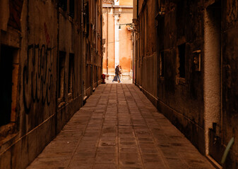 Obraz premium Venice, Italy - February 04, 2024: Narrow alleyway in Venice, Italy, featuring cobblestone pavement and graffiti on old buildings with a couple walking in the distance