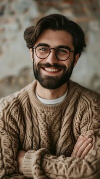 Warm smile radiates from bearded man. Cropped hair, round glasses, soft knit sweater. Arms crossed, relaxed posture against textured wall. Natural lighting enhances his friendly expression